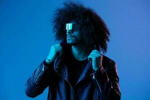 Portrait of fashion man with curly hair with stylish glasses on blue background multinational, colored light, black leather jacket trend, modern concept. photo