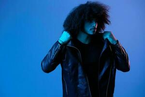 Portrait of fashion man with curly hair on blue background multinational, colored light, black leather jacket trend, modern concept. photo