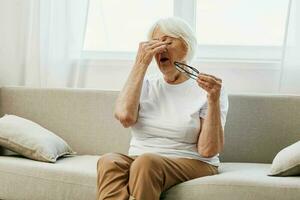 Elderly woman severe eye pain sitting on the couch, health problems in old age, poor quality of life. A grandmother with gray hair holds on to her sore eyes with glasses in her hand. photo