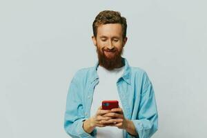 Man hipster with phone in hand blogger smile on gray background in blue shirt and white t-shirt, talking on the phone and selfies photo