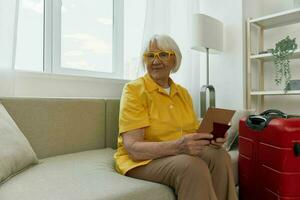 Happy senior woman with passport and travel ticket packed a red suitcase, vacation and health care. Smiling old woman joyfully sitting on the sofa before the trip raised her hands up in joy. photo