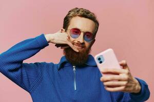 Portrait of a redheaded man with phone in hand taking selfies and photos on his phone with a smile on a pink background, blogger