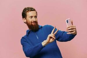 retrato de un pelirrojo hombre con teléfono en mano tomando selfies y fotos en su teléfono con un sonrisa en un rosado fondo, blogger