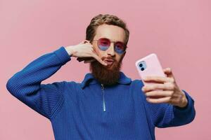 Portrait of a redheaded man with phone in hand taking selfies and photos on his phone with a smile on a pink background, blogger