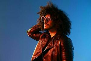 Portrait of a stylish man with curly hair with glasses smile on blue background multinational, color light, black leather jacket trend, modern concept. photo