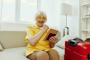 Happy senior woman with passport and travel ticket packed a red suitcase, vacation and health care. Smiling old woman joyfully sitting on the sofa before the trip raised her hands up in joy. photo