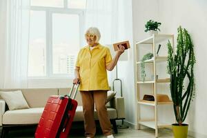 contento mayor mujer con pasaporte y viaje boleto lleno un rojo maleta, vacaciones y salud cuidado. sonriente antiguo mujer alegremente soportes en el casa antes de el viaje. foto