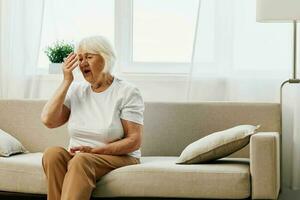 Elderly woman severe pain in the head sitting on the couch, health problems in old age, poor quality of life. Grandmother with gray hair holding her head, migraine and high blood pressure. photo