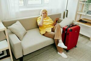 Happy senior woman with passport and travel ticket packed a red suitcase, vacation and health care. Smiling old woman joyfully sitting on the sofa before the trip raised her hands up in joy. photo