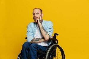 A man in a wheelchair problems with the musculoskeletal system looks at the camera shows a finger on, with tattoos on his hands sits on a yellow studio background, health concept man with disabilities photo