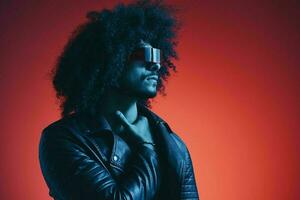 Portrait of fashion man with curly hair on red background with stylish glasses, multicultural, colored light, black leather jacket trend, modern concept. photo