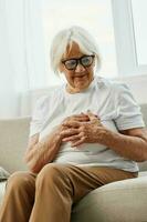 Elderly woman severe chest pain sitting on the sofa, health problems in old age, poor quality of life. Grandmother with gray hair holds her chest with her hands, women's health, breast cancer. photo