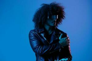 Portrait of fashion man with curly hair with stylish glasses on blue background multinational, colored light, black leather jacket trend, modern concept. photo