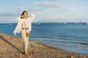 A woman happy with her eyes closed in the sun walking on the beach with her arms spread out against the ocean with a smile, flying hair, tanned skin, relaxation, a trip to the ocean and freedom. photo