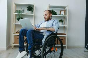 un hombre en un silla de ruedas persona de libre dedicación trabajos a un ordenador portátil a hogar, trabajo en línea, social redes y puesta en marcha, Copiar espacio, integración dentro sociedad, concepto de salud discapacitado persona, real persona de cerca foto