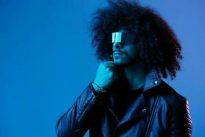 Portrait of fashion man with curly hair with stylish glasses on blue background multinational, colored light, black leather jacket trend, modern concept. photo