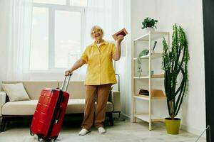 Happy senior woman with passport and travel ticket packed a red suitcase, vacation and health care. Smiling old woman joyfully stands in the house before the trip. photo