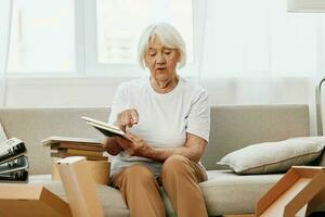 elderly woman sits on a sofa at home with boxes. collecting things with memories albums with photos and photo frames moving to a new place cleaning things and a happy smile. Lifestyle retirement.