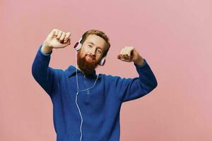 retrato de un pelirrojo hombre vistiendo auriculares sonriente y baile, escuchando a música en un rosado antecedentes. un hipster con un barba. foto