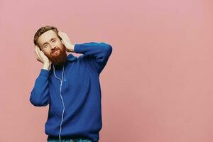 retrato de un pelirrojo hombre vistiendo auriculares sonriente y baile, escuchando a música en un rosado antecedentes. un hipster con un barba. foto