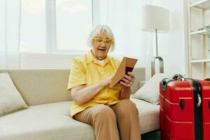 Happy senior woman with passport and travel ticket packed a red suitcase, vacation and health care. Smiling old woman joyfully sitting on the sofa before the trip raised her hands up in joy. photo