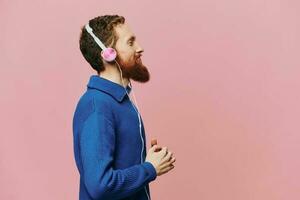 retrato de un pelirrojo hombre vistiendo auriculares sonriente y baile, escuchando a música en un rosado antecedentes. un hipster con un barba. foto