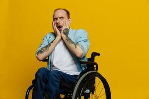 A man in a wheelchair surprised, with tattoos on his arms sits on a yellow studio background, the concept of health is a person with disabilities photo