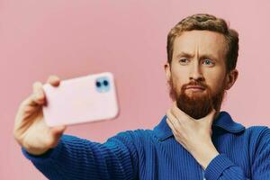 retrato de un pelirrojo hombre con teléfono en mano tomando selfies y fotos en su teléfono con un sonrisa en un rosado fondo, blogger