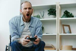 A man in a wheelchair gamer plays games with a joystick in his hands at home, anger and sadness from losing, copy space, with tattoos on his hands, health concept man with disabilities, real person photo