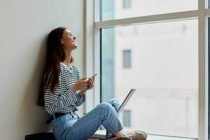 Happy woman sitting on the floor by the windows with a laptop on her lap and holding her phone, a view of the big city, the concept of work and learning online photo