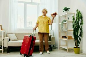 contento mayor mujer con pasaporte y viaje boleto lleno un rojo maleta, vacaciones y salud cuidado. sonriente antiguo mujer alegremente soportes en el casa antes de el viaje. foto