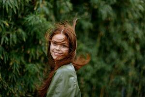 retrato de un hermosa joven niña en el ciudad con un hermosa sonrisa y rojo pelo en un verde impermeable en el ciudad en contra un antecedentes de bambú en primavera, estilo de vida en el ciudad foto