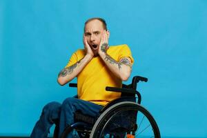 un hombre en un silla de ruedas sorprendido y abierto boca en un camiseta con tatuajes en su brazos se sienta en un azul estudio fondo, un lleno vida, un real persona foto