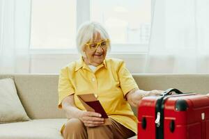 Happy senior woman with passport and travel ticket packed a red suitcase, vacation and health care. Smiling old woman joyfully sitting on the sofa before the trip raised her hands up in joy. photo