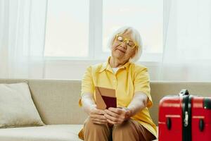 Happy senior woman with passport and travel ticket packed a red suitcase, vacation and health care. Smiling old woman joyfully sitting on the sofa before the trip raised her hands up in joy. photo