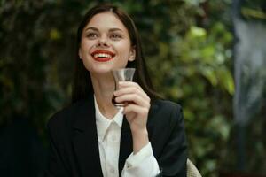 woman with red lips smile with teeth drinks tea in a cafe from a Turkish glass mug in a white shirt and black fashion jacket on the street, summer travel, vacation in the city photo