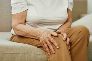 Elderly woman severe pain in her leg sitting on the couch, health problems in old age, poor quality of life. Grandmother with gray hair holds on to her sore knee, problems with joints and ligaments. photo