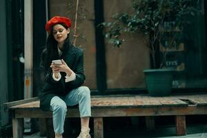 Stylish woman tourist in stylish clothes in a jacket and red beret sitting outside with phone in hand, cell phone in travel, French style, cinematic color. photo