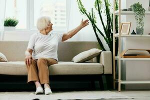 Elderly woman sits on sofa at home, bright spacious interior in old age smile, lifestyle. Grandmother with gray hair in a white T-shirt and beige trousers. photo