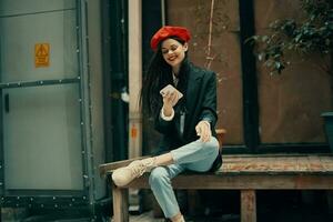 Stylish woman tourist in stylish clothes in a jacket and red beret sitting outside with phone in hand, cell phone in travel, French style, cinematic color. photo