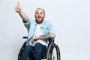 A man in a wheelchair looks at the camera shows a thumbs up, happiness, with tattoos on his hands sits on a gray studio background, health concept man with disabilities photo