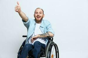 un hombre en un silla de ruedas mira a el cámara muestra un pulgares arriba, felicidad, con tatuajes en su manos se sienta en un gris estudio fondo, salud concepto hombre con discapacidades foto