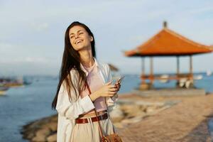 Woman smile blogger freelancer with phone in hand on vacation walking on the beach by the ocean in Bali, happy travel and vacation, mobile communications, internet online photo