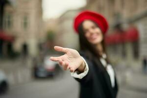 Moda mujer sonrisa con dientes en pie en el calle en frente de el ciudad turista seguir yo elegante ropa con rojo labios y rojo boina, viajar, cinematográfico color, retro Clásico estilo, urbano moda. foto