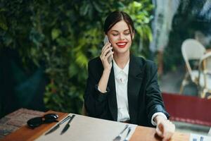 un mujer blogger turista se sienta en un café a un mesa con un teléfono en su manos hablando en el teléfono, móvil comunicaciones y el Internet en un primavera viaje, negocio llamar, Lanza libre trabajo en línea, sonrisa foto