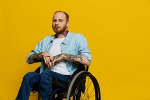 A man in a wheelchair problems with the musculoskeletal system looks at the camera thoughtfully with tattoos on his arms sits on a yellow studio background, health concept a person with disabilities photo