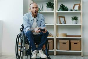 A man in a wheelchair gamer plays games with a joystick in his hands at home, surprise open mouth, copy space, with tattoos on his hands, health concept man with disabilities, real person photo