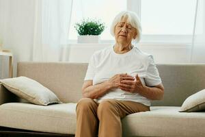 Elderly woman severe pain in the heart sitting on the couch, health problems in old age, poor quality life. Grandmother with gray hair holding on her chest, heart attack and stroke, pressure problem. photo