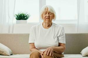 Elderly woman severe pain in her leg sitting on the couch, health problems in old age, poor quality of life. Grandmother with gray hair holds on to her sore knee, problems with joints and ligaments. photo