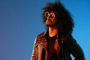 Portrait of a stylish man with curly hair with glasses smile on blue background multinational, color light, black leather jacket trend, modern concept. photo